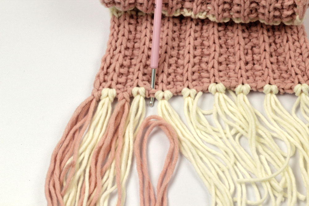 Schal stricken - Mit der Häkelnadel stichst du von vorne nach hinten in die Masche ein, um dann das auf die Hälfte geknickte Fransenbüschel einzuknüpfen.