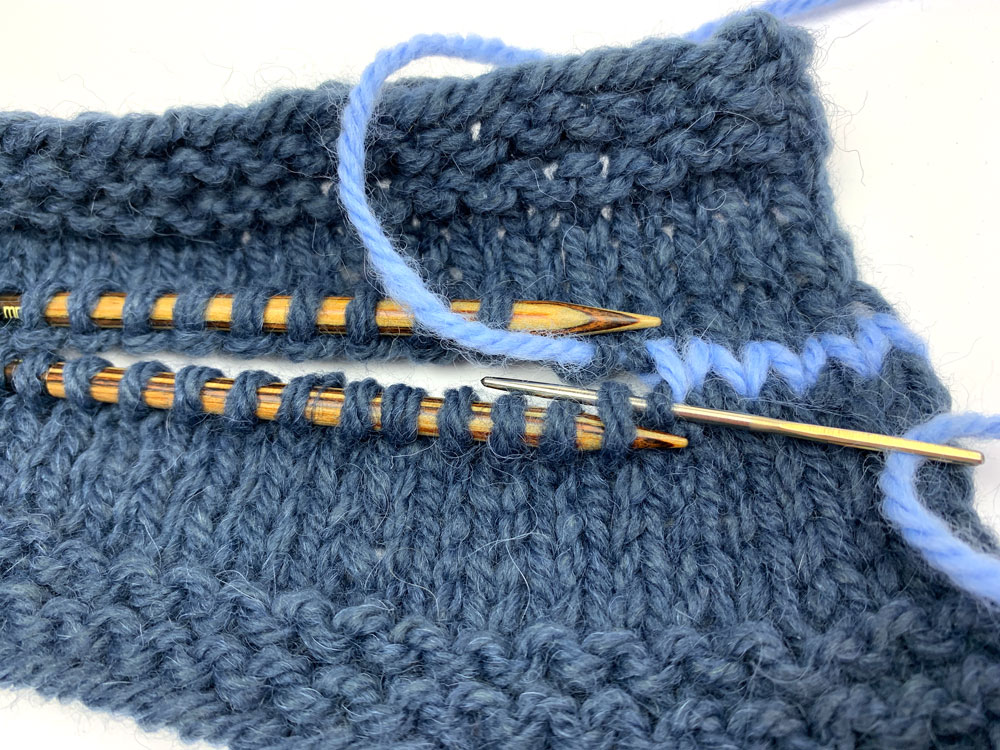 Schritt 6: Auf der unteren Stricknadel schiebst du die Nadel an der Ausstichstelle des Fadens von vorne in die Masche neben der Nadelspitze und gleich im Anschlussvon hinten durch die Masche auf der Stricknadel.