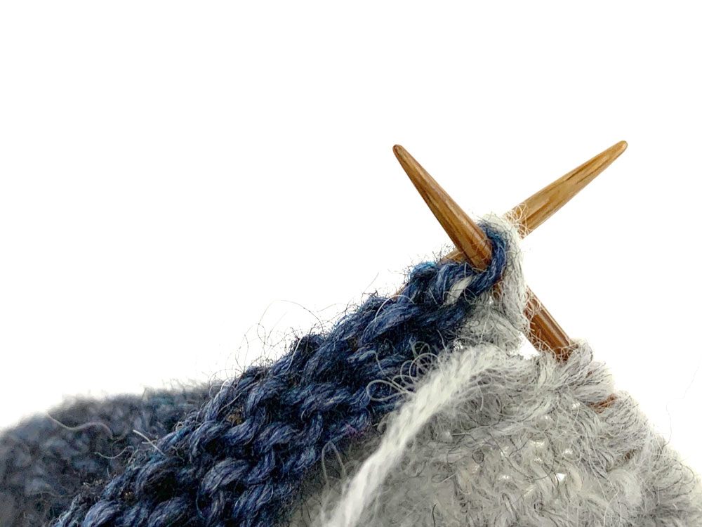 Die letzte Masche von Nadel 3 wird auf Nadel 2 geschoben und mit dem Umschlag links zusammengestrickt.