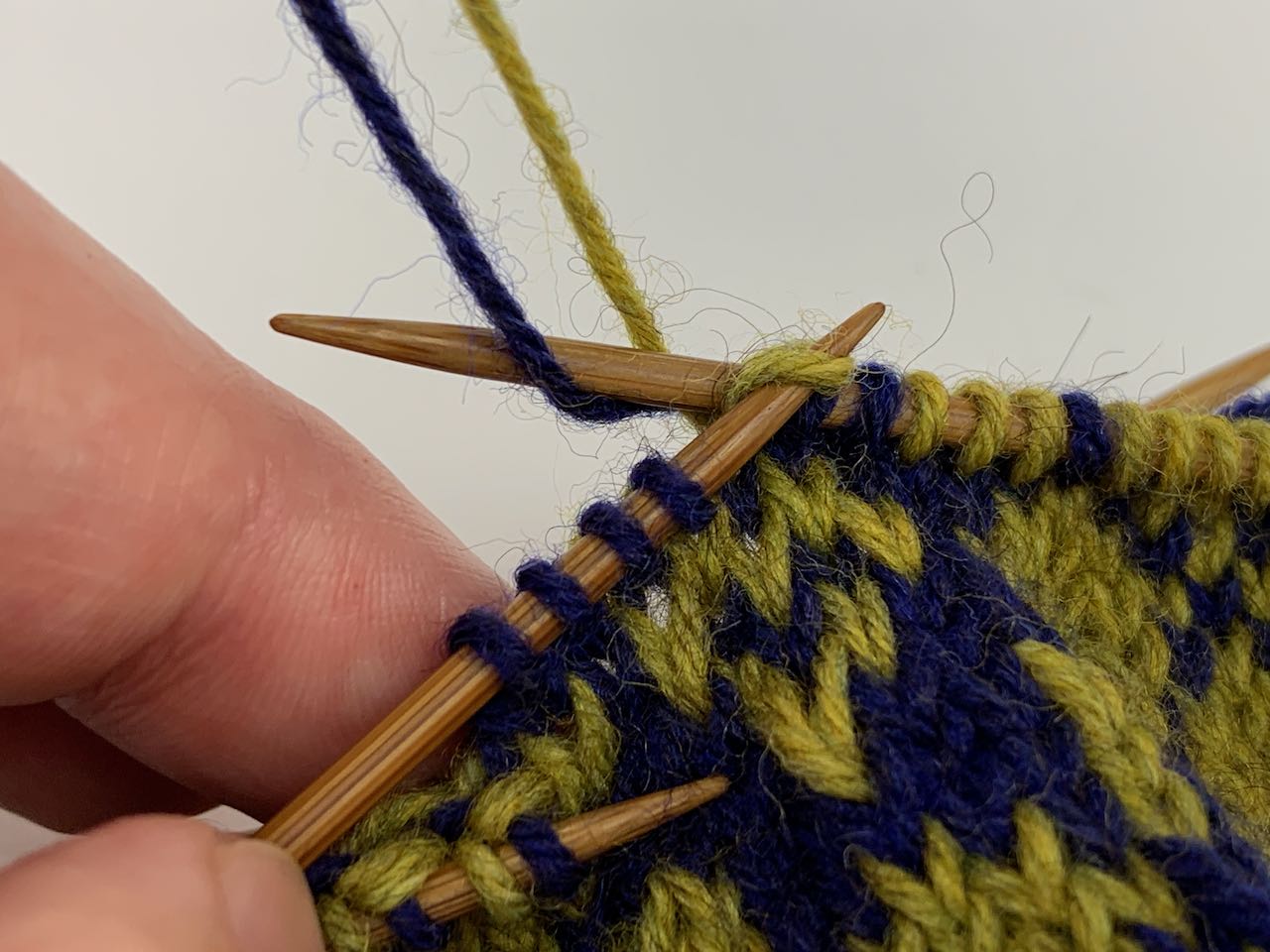 Das Verweben von Spannfäden beim Jacquardmuster stricken. Du strickst den hinteren Faden und verwebst den vorderen Faden – Lege den vorderen Faden über die Nadel mit der Masche, die gleich abgestrickt wird.